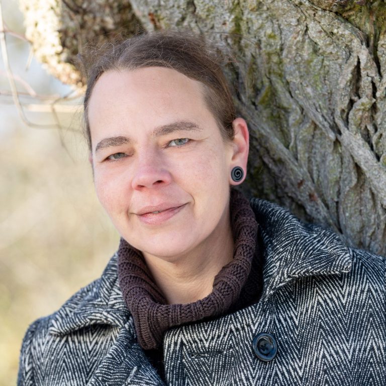 Frau mit Dutt und Ohrringen, trägt einen grauen Mantel, steht vor einem Baum.